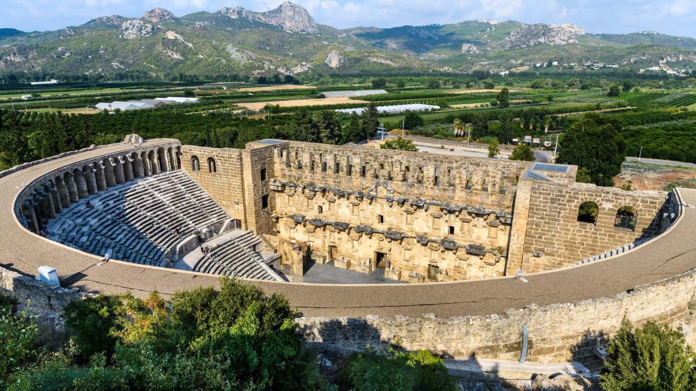 An aerial view of an ancient stone amphitheater surrounded by mountains and greenery.