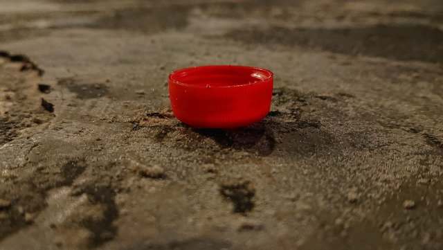 A red plastic bottle cap sits on a textured, dark surface with visible dirt and debris around it.