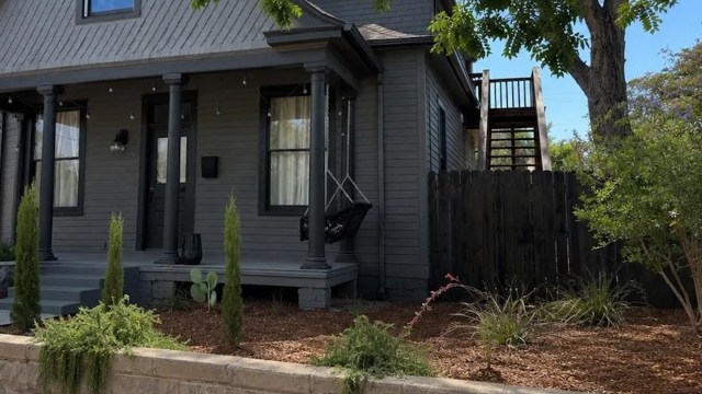 A modern gray house with a front porch, swing chair, and landscaped yard featuring shrubs and a wooden fence.