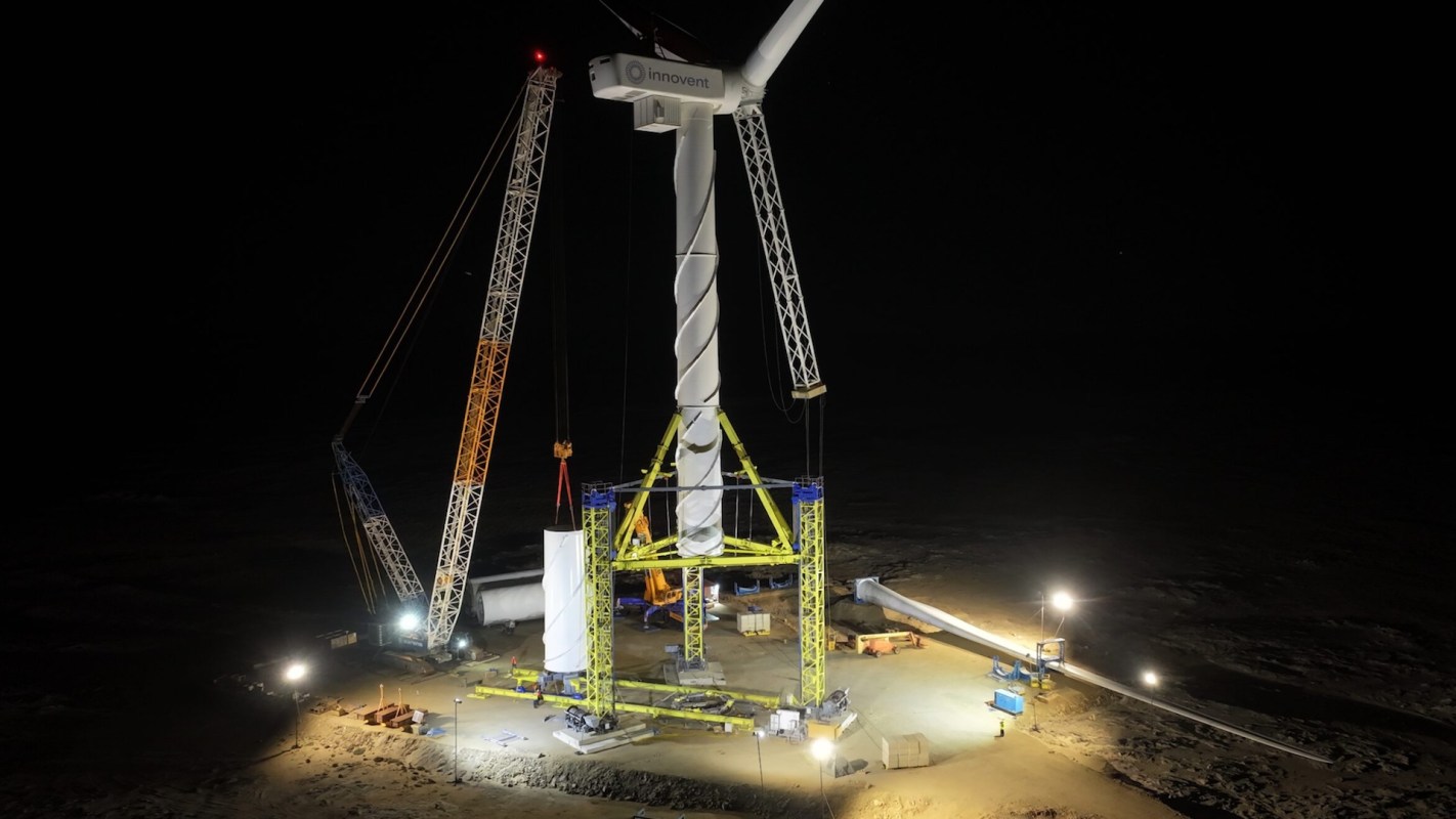 A wind turbine installation site at night, featuring cranes and bright lights illuminating the area.