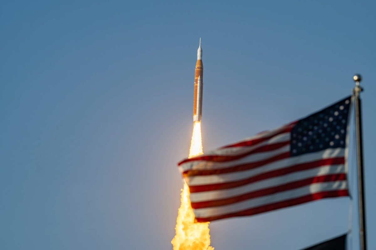 A rocket launches against a clear blue sky, with a waving American flag in the foreground.