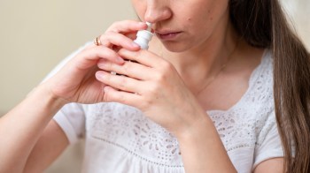 A woman with long hair holds a nasal spray bottle close to her nose, preparing to use it.