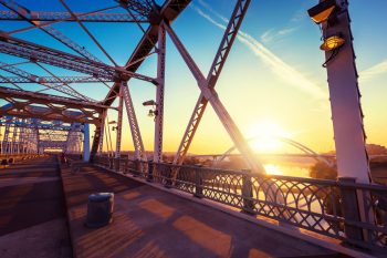 A metal bridge with intricate supports at sunset, reflecting over a river with city buildings in the background.