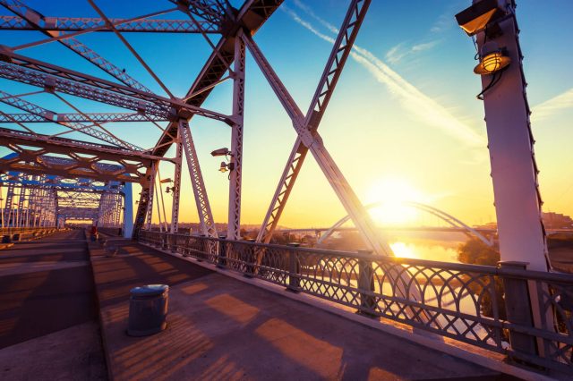 A metal bridge with intricate supports at sunset, reflecting over a river with city buildings in the background.