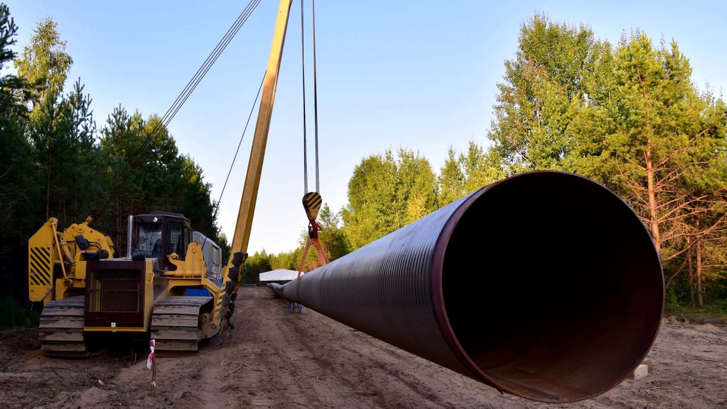 A large pipe is being lifted by a crane near a forested area with machinery in the foreground.