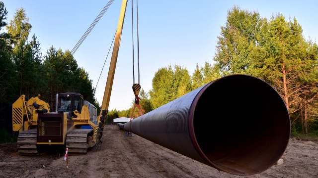 A large pipe is being lifted by a crane near a forested area with machinery in the foreground.