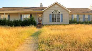 An abandoned single-story house with blue shutters surrounded by unkempt, dry grass on a sunny day.