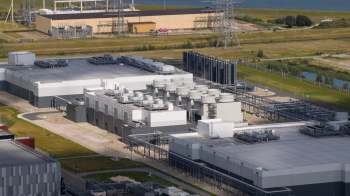 An aerial view of an industrial complex with large buildings, cooling towers, and power lines in a grassy landscape.