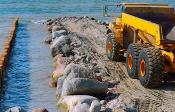 A yellow dump truck on a sandy shore near water, alongside a rocky barrier in the background.