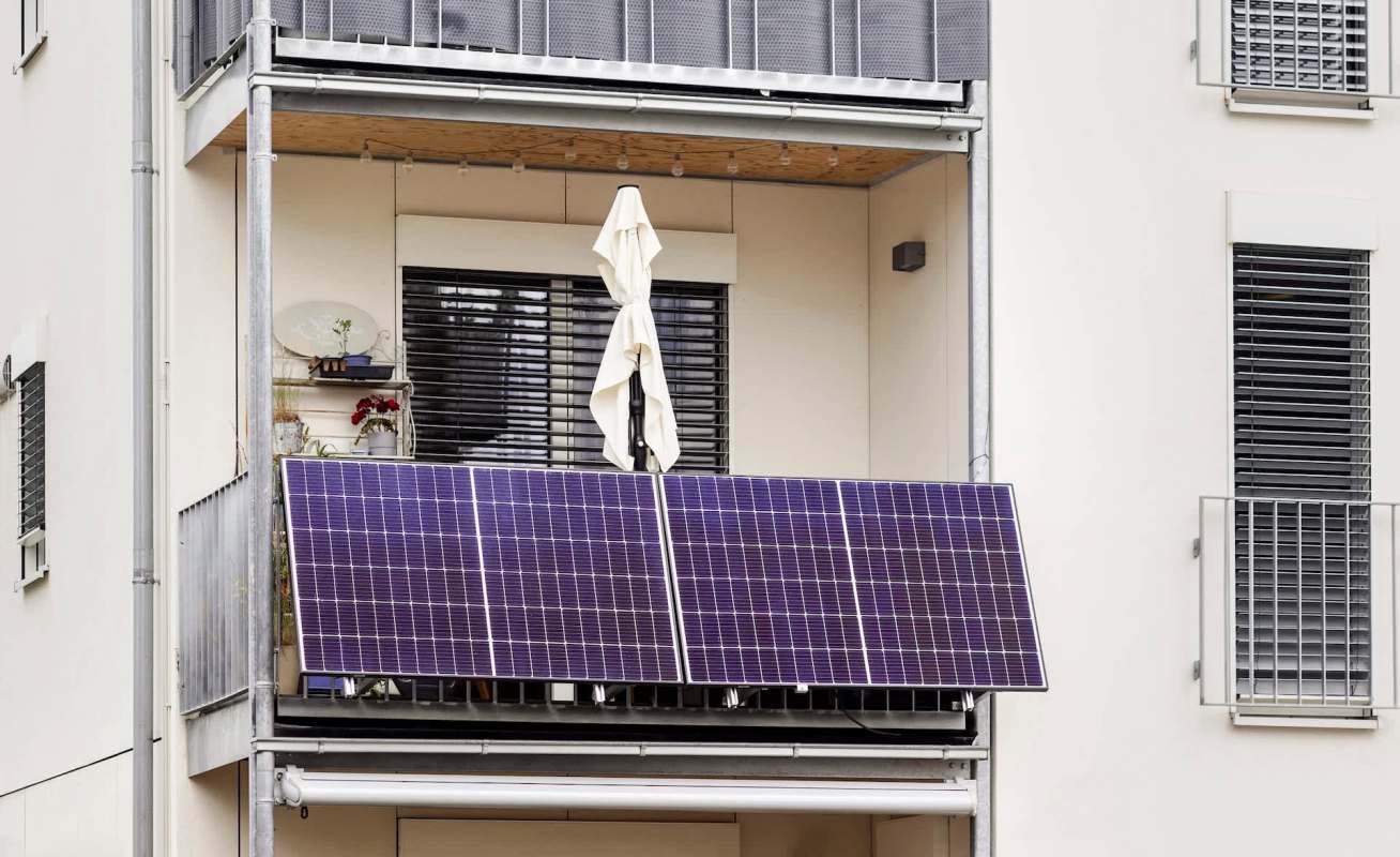 A balcony featuring solar panels, a white umbrella, and potted plants against an apartment building backdrop.
