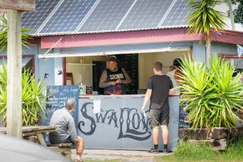 A tattooed vendor serves customers in a tropical setting at a food stall named "Swallow."