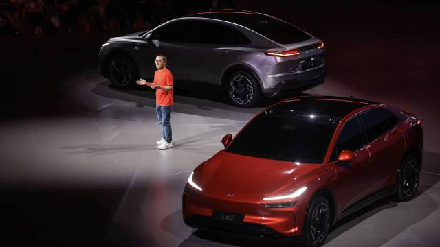 A presenter gesturing near two modern electric SUVs, one red and one silver, at a vehicle launch event.