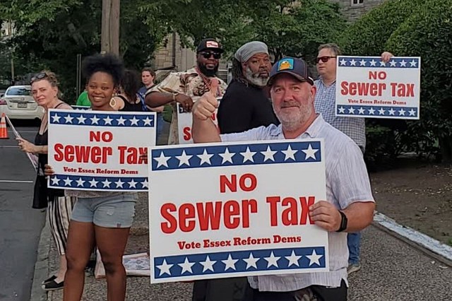 A group of people protesting with signs that read "No Sewer Tax" in an outdoor setting.