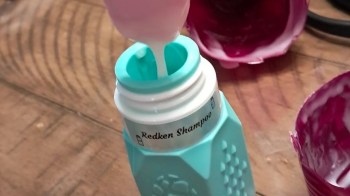 A close-up of a teal shampoo bottle being filled with creamy pink shampoo, set on a wooden surface.