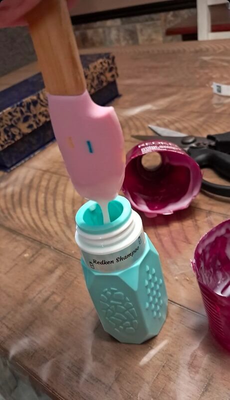 A close-up of a teal shampoo bottle being filled with creamy pink shampoo, set on a wooden surface.