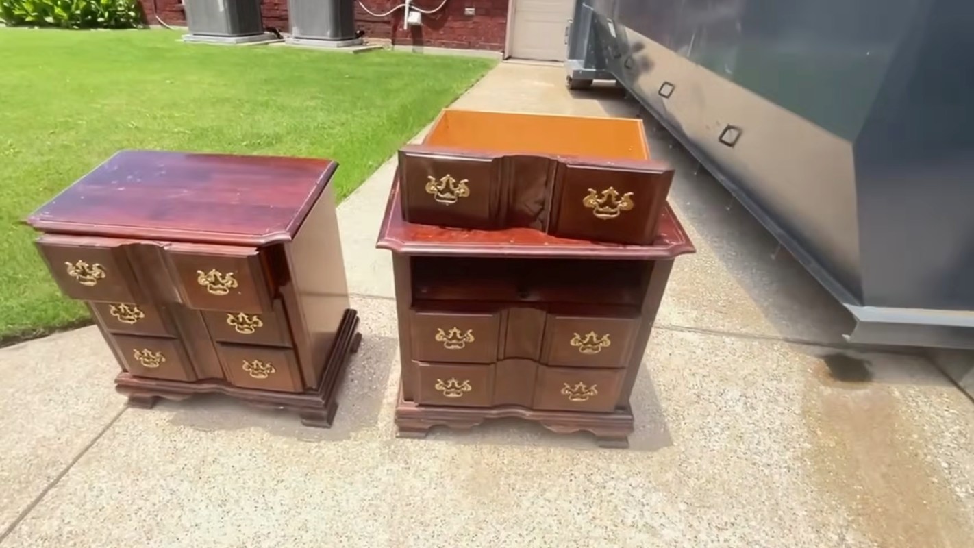 Two wooden nightstands with brass handles displayed on a concrete surface, next to a dumpster in front of a house.