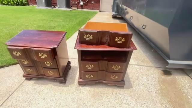 Two wooden nightstands with brass handles displayed on a concrete surface, next to a dumpster in front of a house.