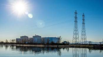 A power plant with cylindrical structures beside a calm waterway, framed by tall power lines under a clear blue sky.