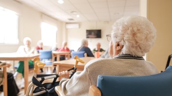 An elderly woman sits in a wheelchair, watching a group of seniors engaged in conversation in a bright room.