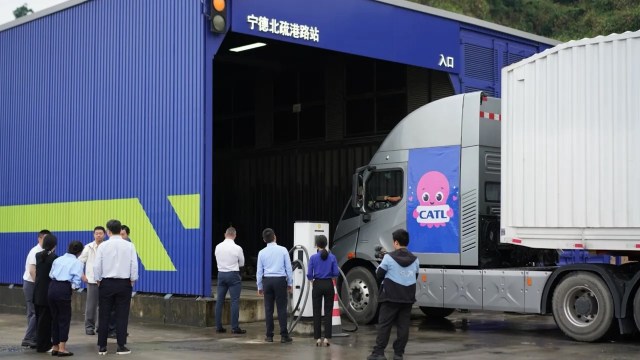 A group of people observes an electric truck charging at a modern station with a blue and green building behind.