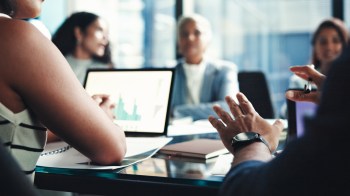 A group of professionals engages in a discussion around a conference table with a laptop displaying graphs.