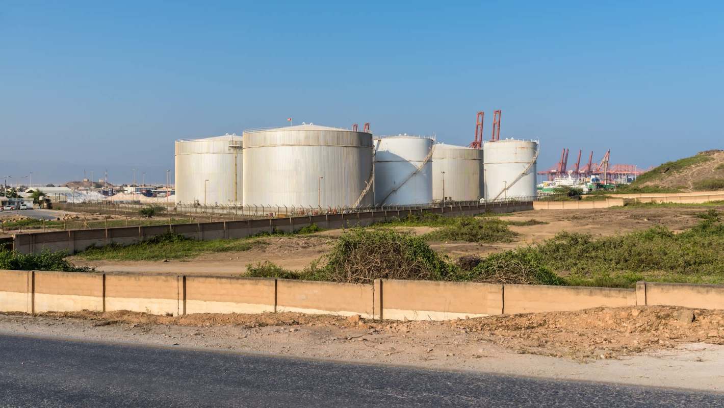 Large industrial oil storage tanks are seen behind a fence, with cranes and shipping containers in the background.
