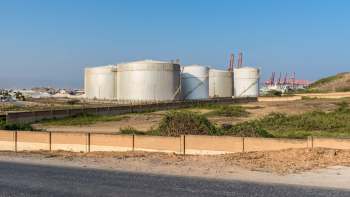 Large industrial oil storage tanks are seen behind a fence, with cranes and shipping containers in the background.