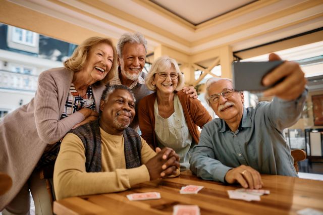 A group of five smiling seniors taking a selfie at a wooden table in a bright, cozy cafe.
