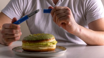 A person holds a blue pen while sat at a table with a green frosted pastry on a plate.