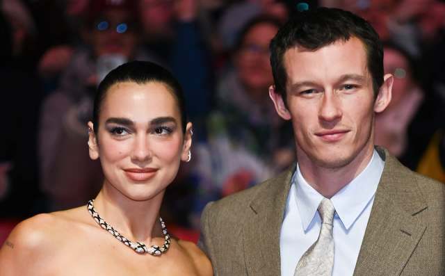 Dua Lipa, in a stylish outfit, poses with her fiance Callum Turner in formal attire, with a crowd in the background.