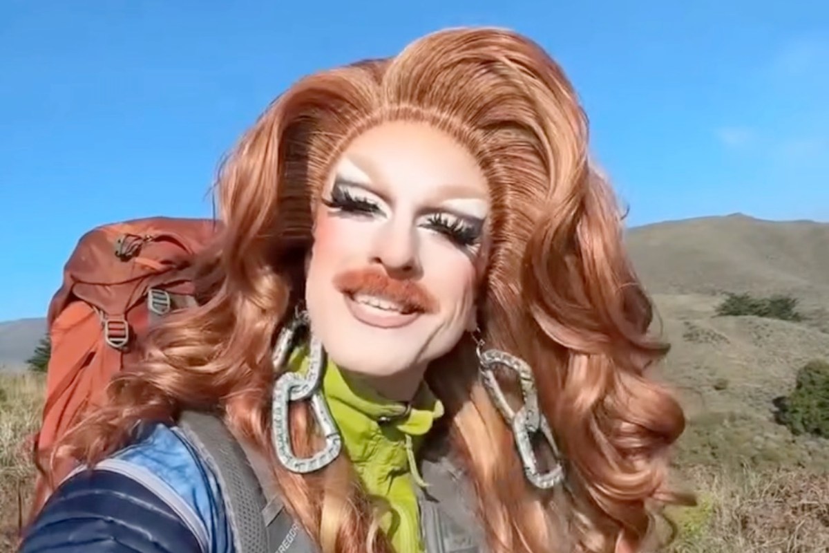 A smiling drag queen with long, styled hair and bold makeup poses outdoors with a backpack and hills in the background.