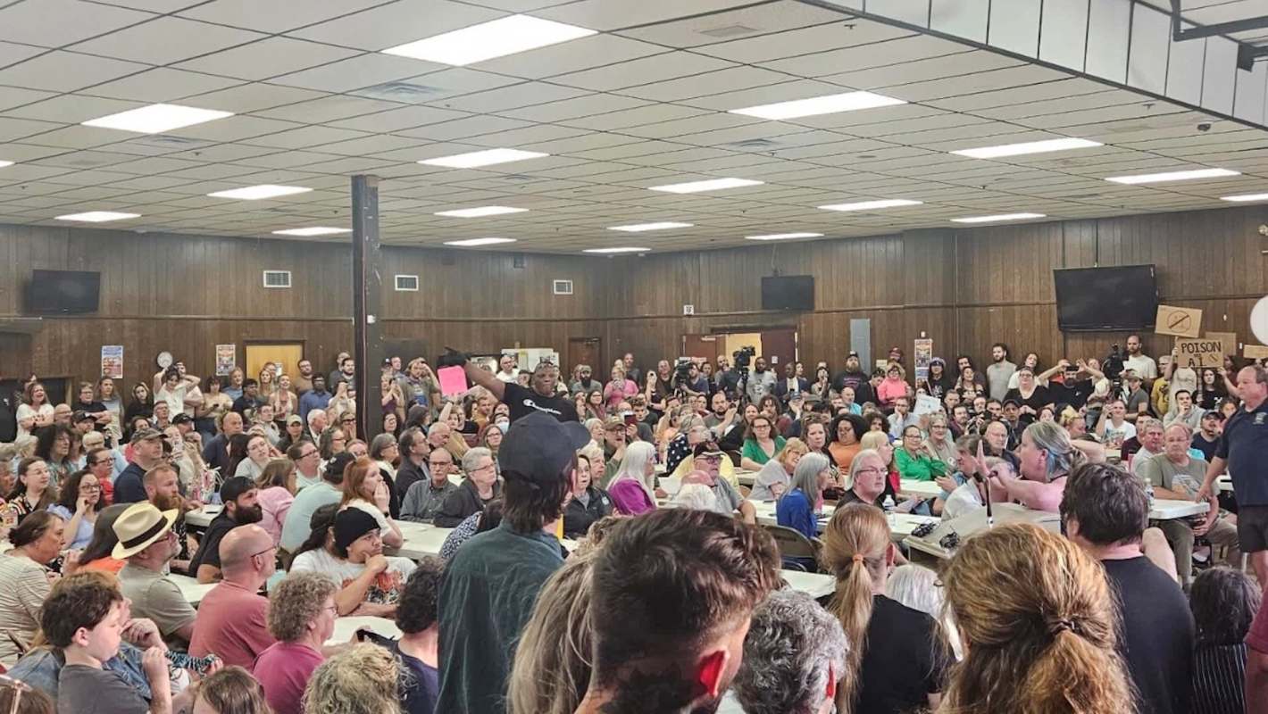 A crowded community meeting with people holding protest signs.