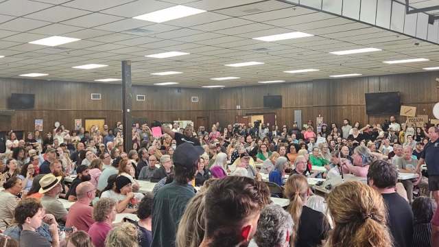 A crowded community meeting with people holding protest signs.
