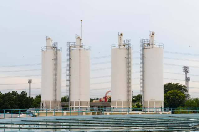 Five white industrial silos stand against a muted sky, with power lines and greenery in the background.