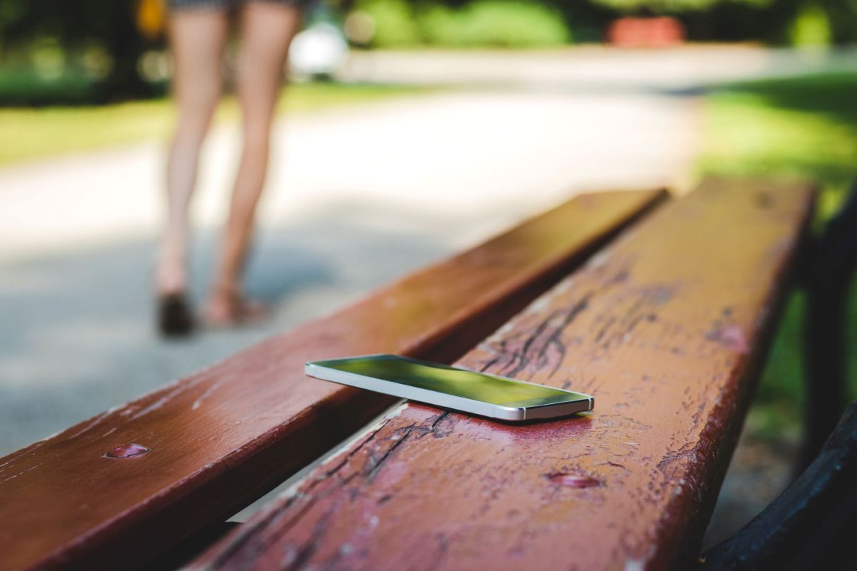 A smartphone rests on a weathered wooden bench, with a blurred figure walking in the background.