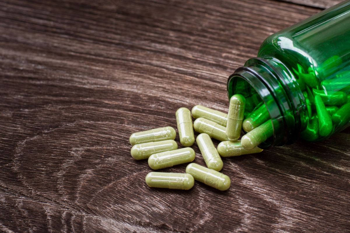 A green bottle tipped over, spilling several light green capsules on a wooden surface.