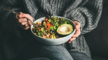 A person in a cozy sweater holds a bowl of colorful salad with avocado, greens, and vegetables.