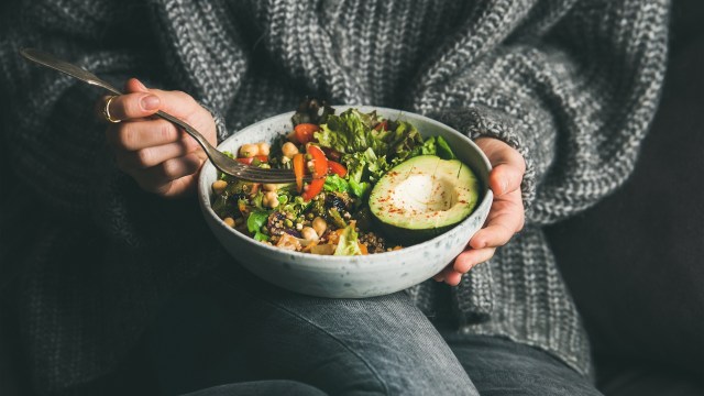 A person in a cozy sweater holds a bowl of colorful salad with avocado, greens, and vegetables.