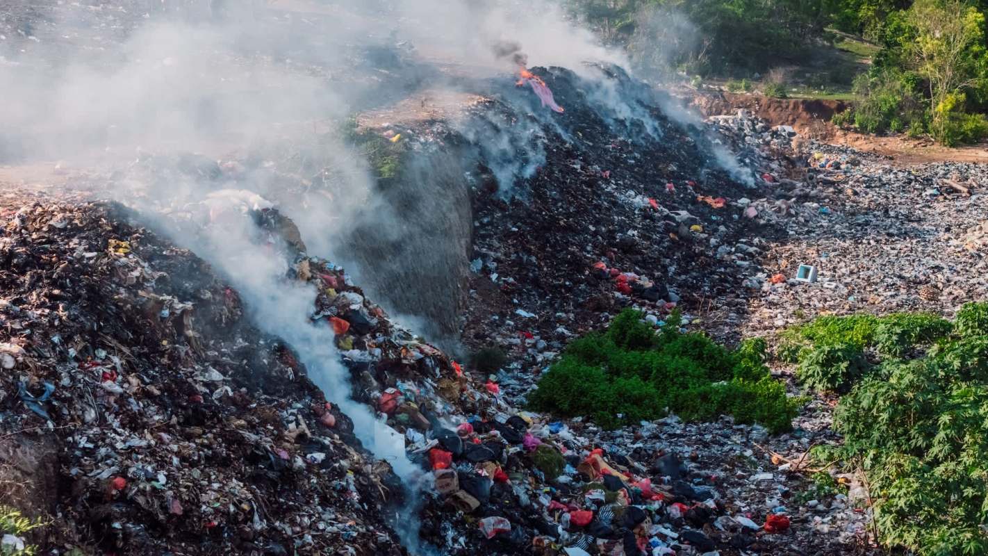 A large landfill emitting smoke with piles of garbage scattered and some greenery nearby.