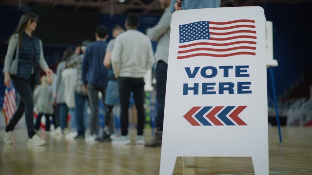A voting location sign with American flags and a line of voters in a gymnasium setting.