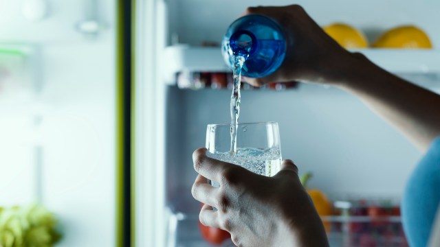 A person pours water from a blue bottle into a glass inside a refrigerator.