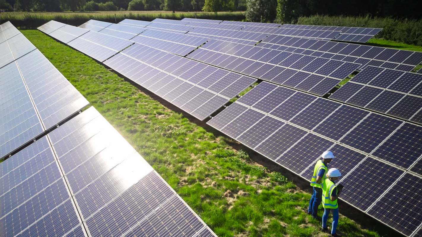 Two workers in helmets inspect solar panels in a green field under bright sunlight.