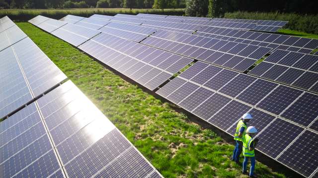 Two workers in helmets inspect solar panels in a green field under bright sunlight.