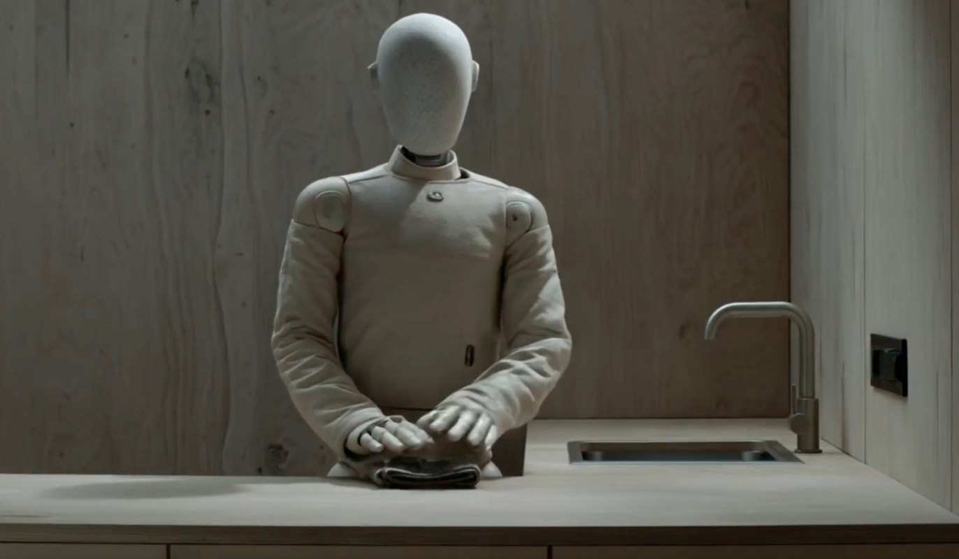 A humanoid robot in a minimalist kitchen folds a towel on a wooden countertop.