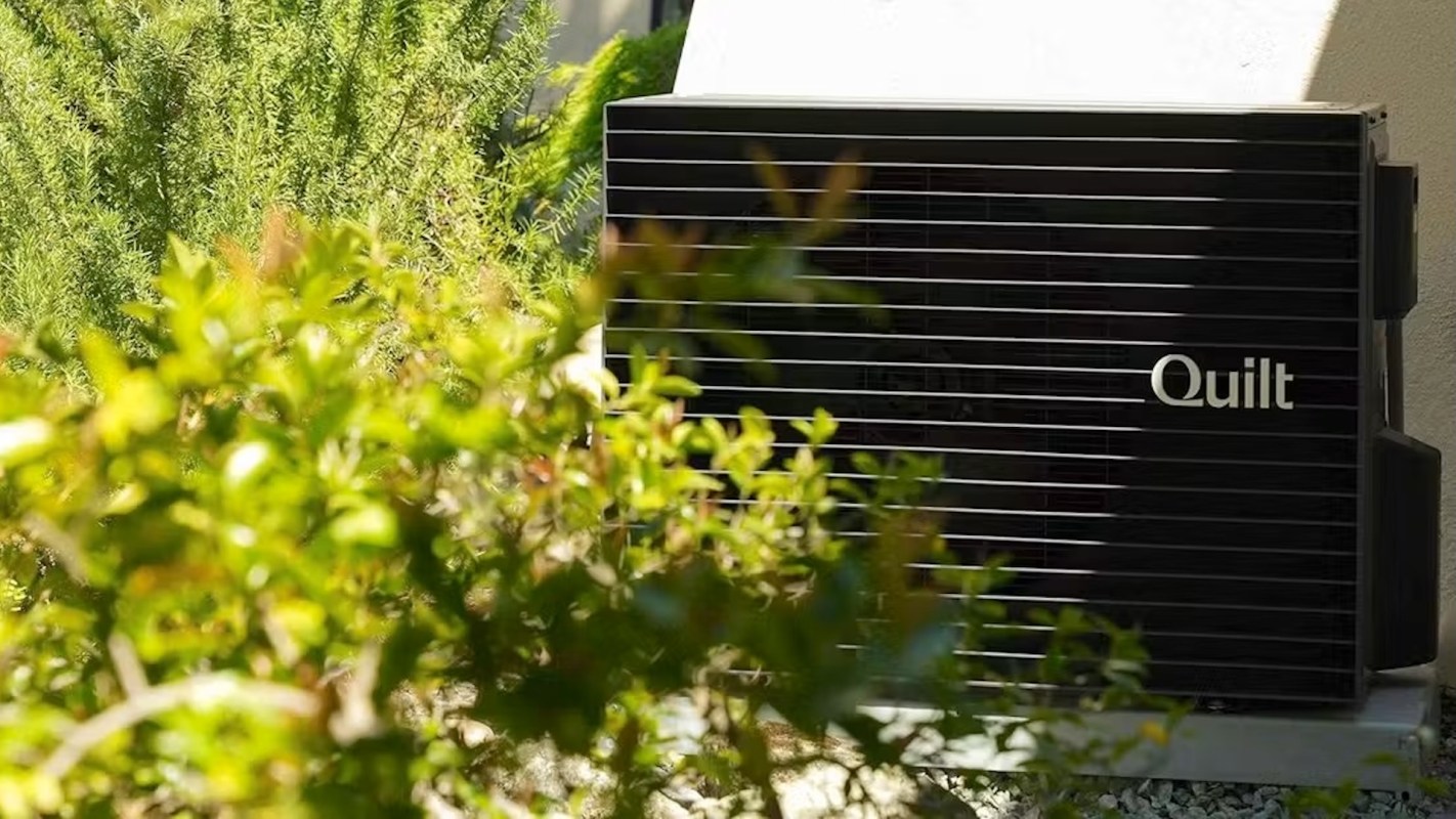 A black outdoor heat pump unit labeled "Quilt" surrounded by lush green plants.