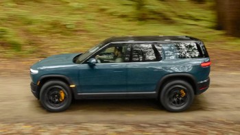 A blue SUV with yellow brake calipers drives along a dirt path surrounded by lush green trees.