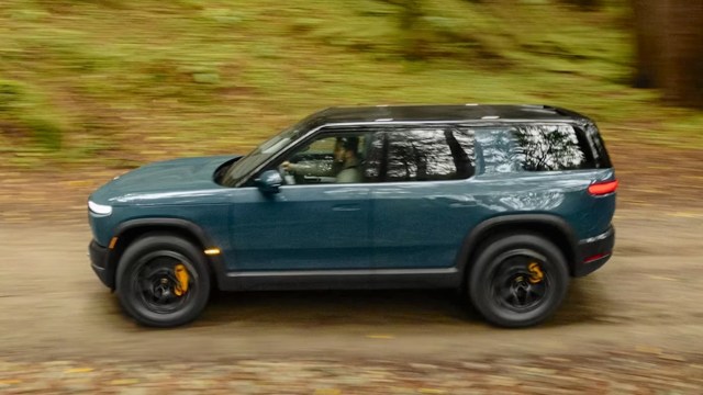A blue SUV with yellow brake calipers drives along a dirt path surrounded by lush green trees.