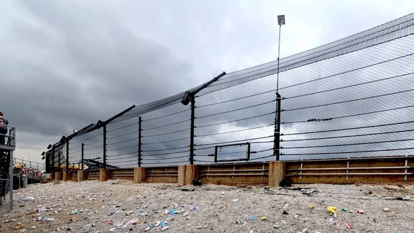 A raceway fence overlooking a litter-strewn area under a cloudy sky, with spectators visible in the background.
