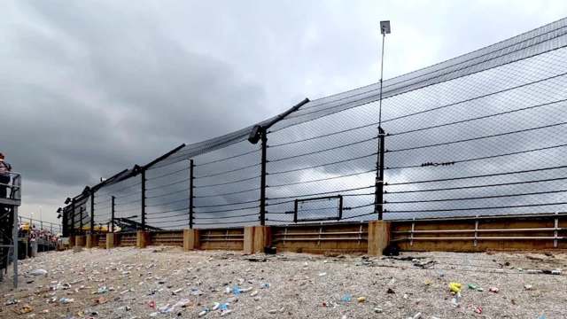 A raceway fence overlooking a litter-strewn area under a cloudy sky, with spectators visible in the background.