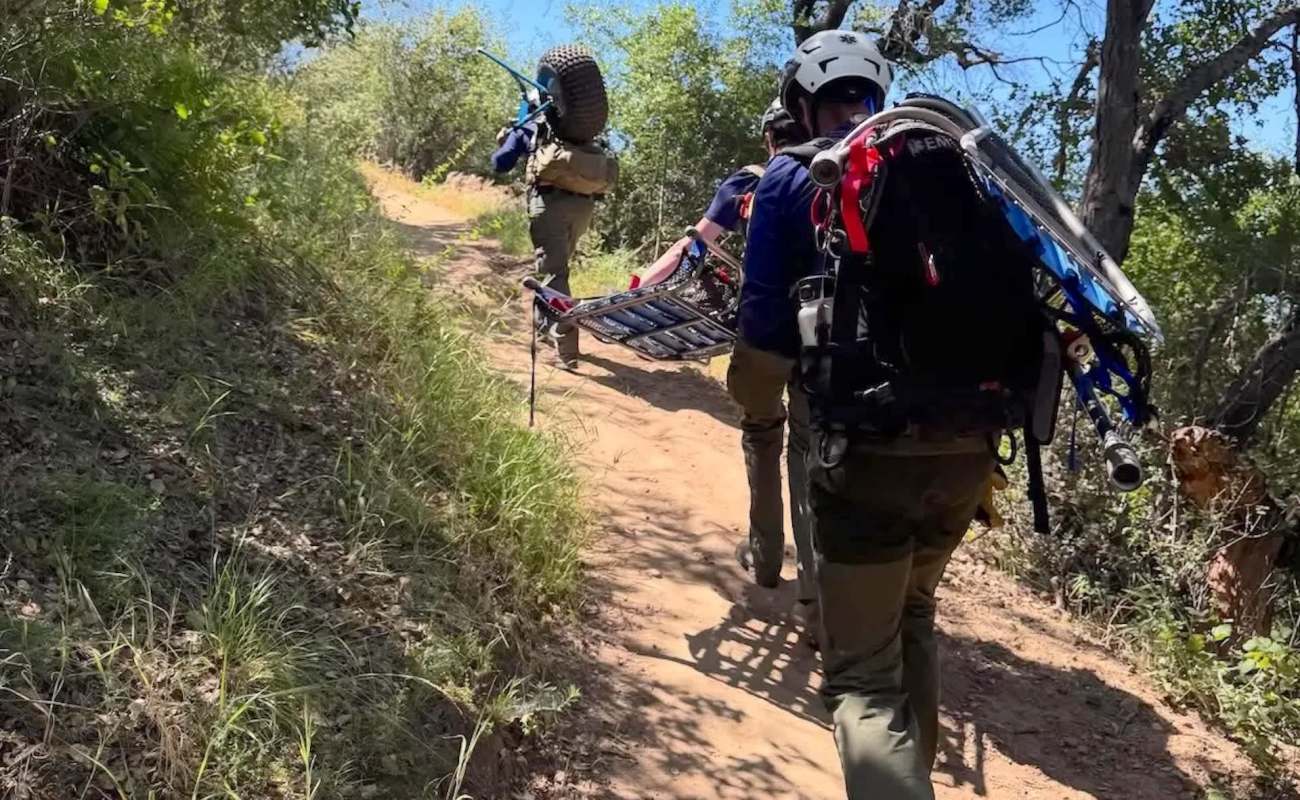 A group of rescuers carries a stretcher along a dirt trail surrounded by lush greenery.